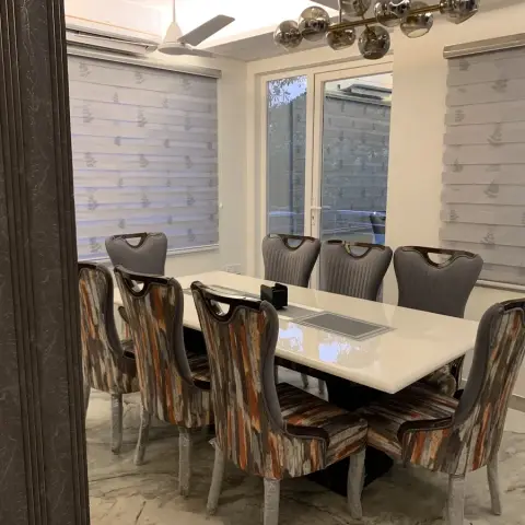 An eight-seater dining room with a white glossy table, patterned wingback dining chairs, a chrome sputnik chandelier and a marble-clad column.