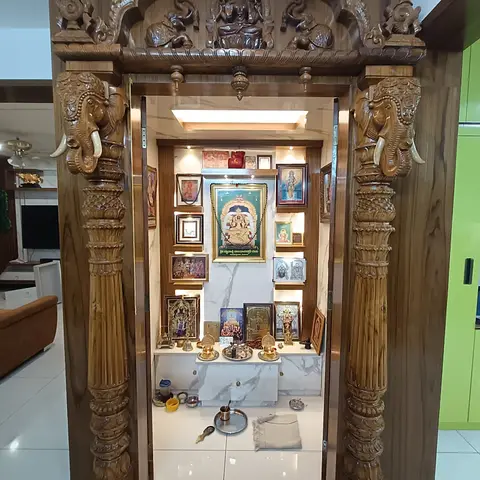 An elaborately hand-carved teak temple unit with twin elephant-head brackets and pillars framing a marble-backed shrine wall lined with multiple deity frames