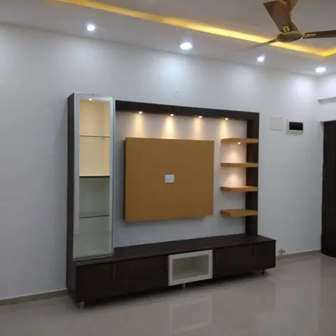 An empty living room wall with a wood-and-glass TV unit and open display shelving beneath a cove-lit false ceiling niche.
