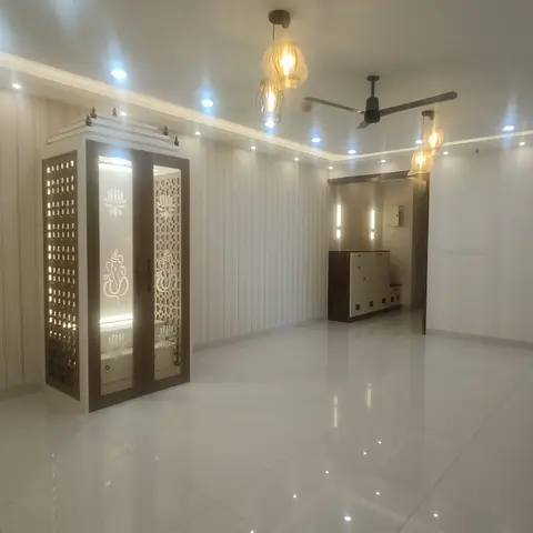 An empty living room with a freestanding carved-wood pooja mandir featuring jali doors with Ganesh and lotus motifs