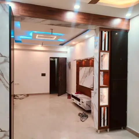 An empty living room with a marble-clad TV unit under a wood-beam ceiling with blue cove lighting, and a marble pillar with black glossy display niches