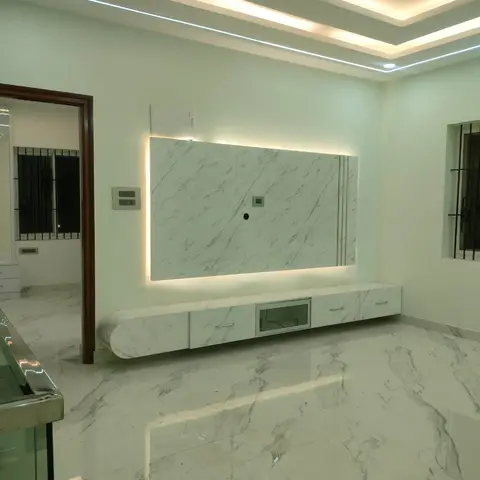 An empty living room with a backlit marble-clad TV wall, matching marble flooring, and a multi-tier cove-lit false ceiling.