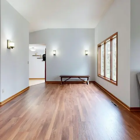 An empty living room with glossy wood flooring, a wood bench beneath a window, a grey sofa, and wall sconces along a vaulted ceiling hallway.