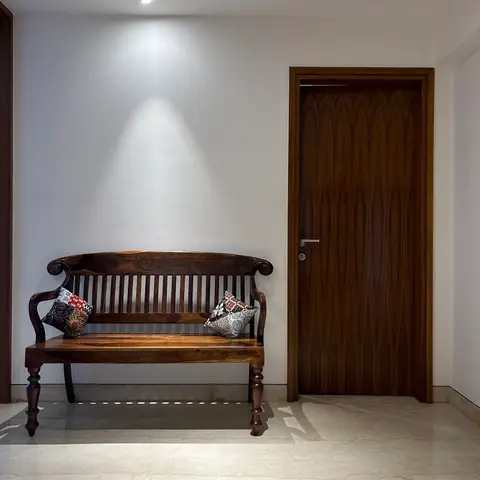 An entrance foyer with a carved wood colonial-style bench beneath a spotlight, beside a vertically fluted teak main door.