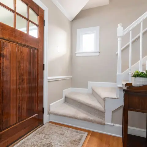 An entry foyer with a stained wood-panel front door, a carpeted staircase, and a dark wood console table with a round mirror.