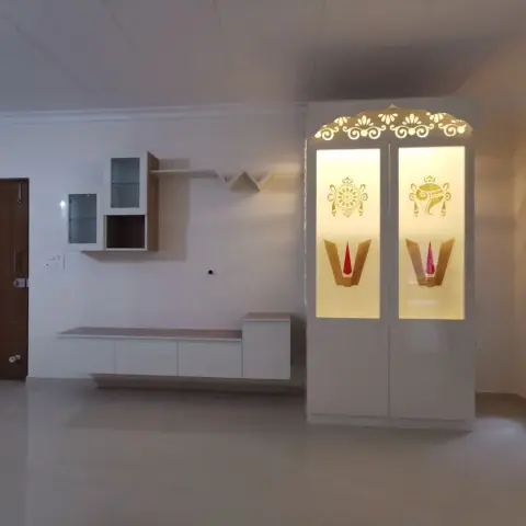 An entryway wall with a backlit white pooja cabinet carved with Om and kalash motifs, beside a floating TV console and open display shelving.