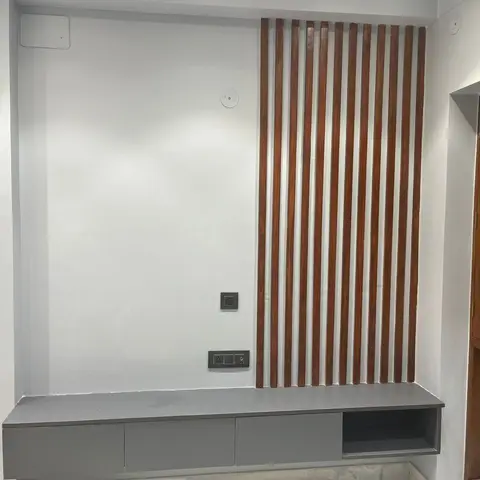 An in-progress TV wall with vertical wood-toned fluted battens beside a plain white wall, above a floating grey low console with a shelf cutout.