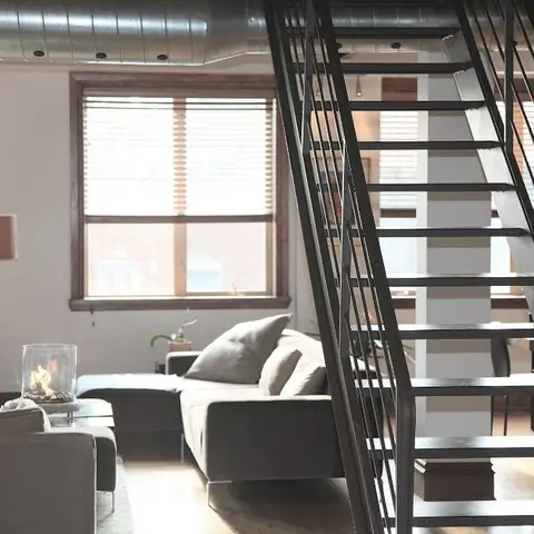 An industrial-style loft living room with exposed ceiling ductwork, a black open-tread metal staircase, and grey sofas around a glass hurricane lantern.