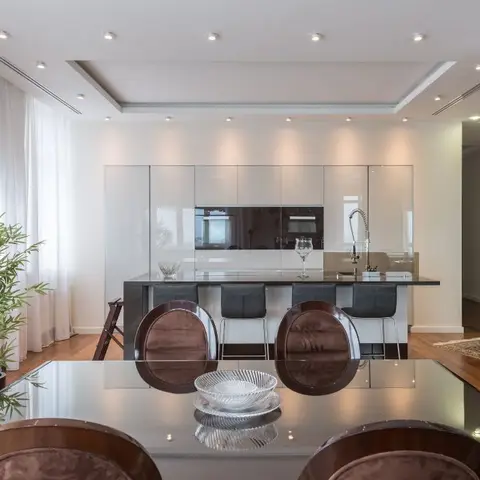 An open dining and kitchen area with a glass dining table, velvet oval-back chairs, and a glossy handleless kitchen island behind.
