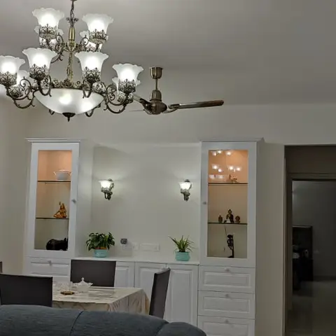 An open dining-kitchen-living space with a scalloped brass chandelier over a dining table, a white glass-front crockery unit with lit display shelves