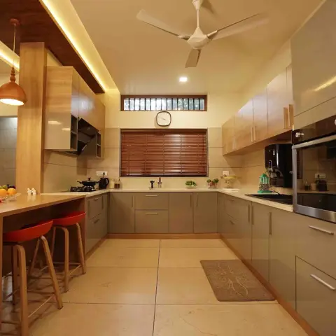 An open kitchen with a wood breakfast counter and red bar stools, twin wood dome pendant lights, a grey glossy L-shaped cabinet run