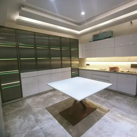 An open kitchen with glossy white cabinetry, a marble backsplash, a fluted glass pantry wall lit with green LED strips, and a sculptural white dining table.