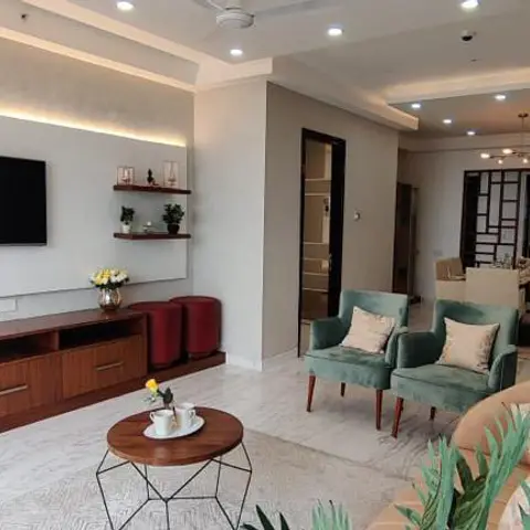 An open living area with a wood-veneer TV console under cove lighting, teal velvet armchairs around a wire-frame coffee table