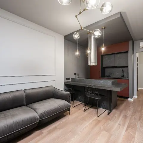 An open living-kitchen space with a dark grey sofa on wood flooring facing a rust-and-charcoal kitchen island lit by a branching brass pendant cluster.