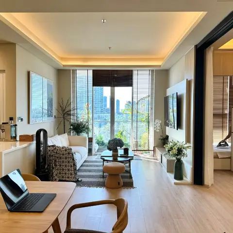 An open-plan apartment showing a dining table, kitchen counter, living room with white sofa and wall-mounted TV, and a bedroom glimpsed through a sliding door