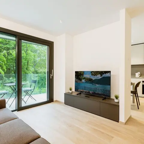 An open-plan apartment shows a living area with a floating TV console, a sliding door onto a balcony with a bistro table, and a white kitchen beyond.