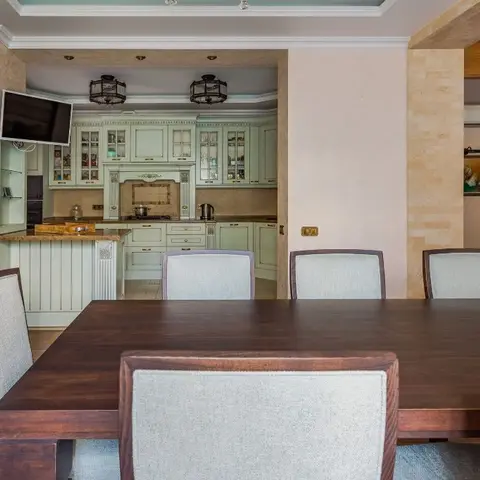An open-plan dining and kitchen with a dark wood dining table for eight in the foreground and a sage-green classic kitchen with glass-front crockery units