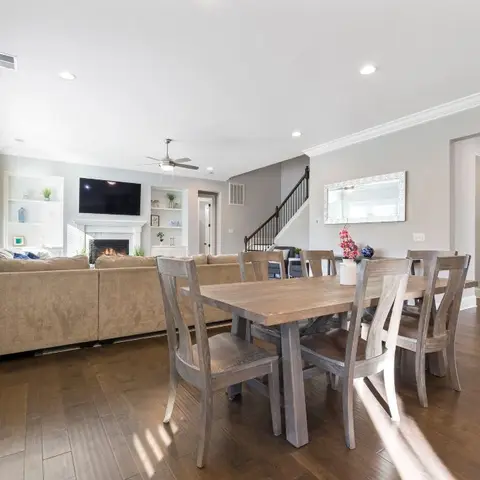 An open-plan dining and living area with a wood dining table, a fireplace with a wall-mounted TV, and a staircase visible in the background.