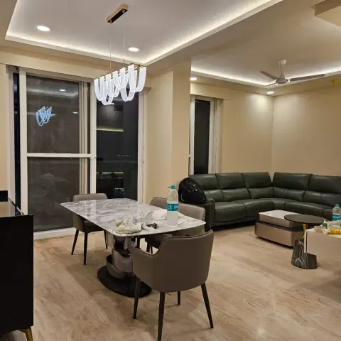 An open-plan dining and living area with a marble dining table, statement pendant light and an olive-green leather sectional, cluttered with everyday items.