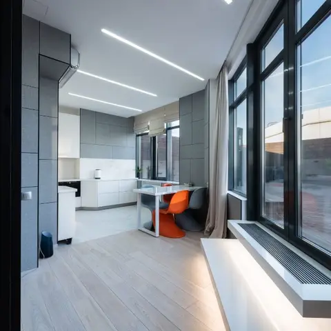 An open-plan dining nook with orange Panton-style chairs at a white table beside floor-to-ceiling windows, a grey-panelled galley kitchen behind.