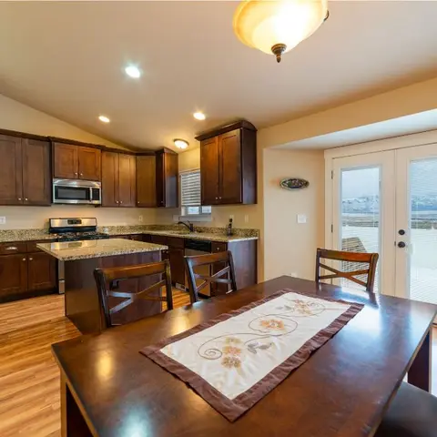 An open-plan dining table with a floral table runner in the foreground, opening onto a dark wood kitchen with an island and French doors leading to a deck.