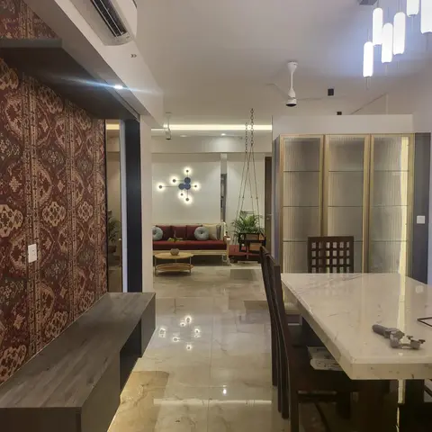 An open-plan dining-to-living view with a white marble dining table and gold-trim glass crockery cabinet in the foreground, leading to a maroon sofa