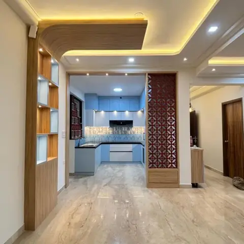 An open-plan entrance flowing past a red carved jaali partition and a backlit display shelf into a powder-blue modular kitchen