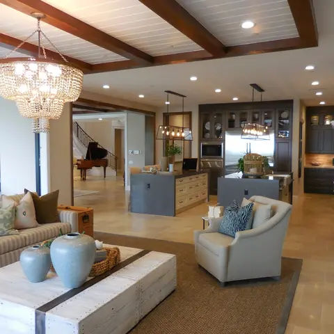 An open-plan great room with a beaded chandelier over a striped sofa in the foreground, two dark kitchen islands under drum pendants