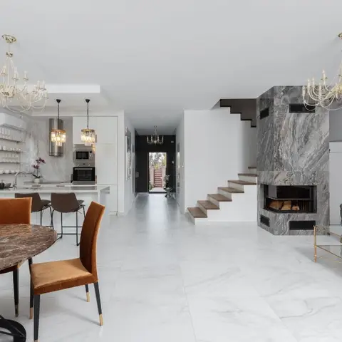 An open-plan ground floor showing a marble-clad fireplace and staircase, a round marble-top dining table under a crystal chandelier