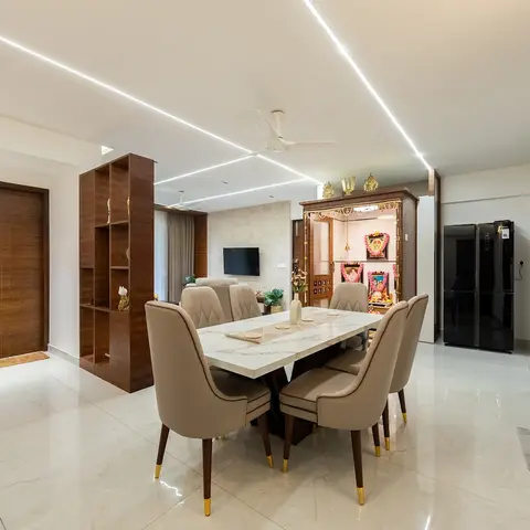 An open-plan home showing a marble dining table, a wood-framed pooja unit, and a living room with sofa and TV, linked by a linear cove-lit ceiling.