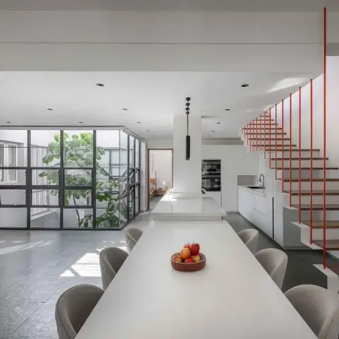 An open-plan kitchen and dining space with a long white island-dining table, built-in wall ovens, a black steel-framed glass wall to an internal courtyard