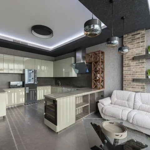 An open-plan kitchen and living area with glossy cream kitchen cabinets and a dark stone island, black cluster pendant lights