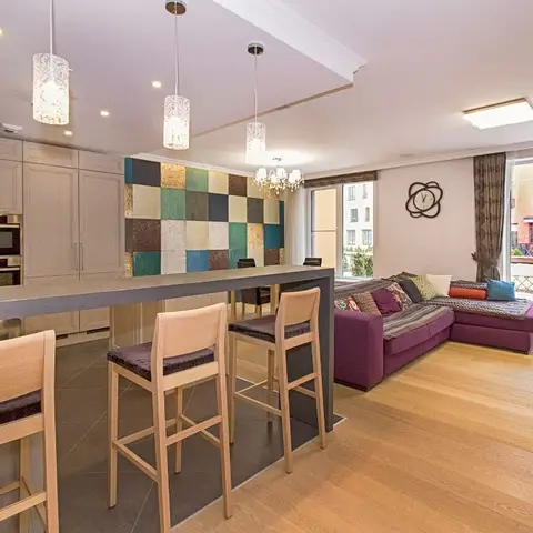 An open-plan kitchen and living space with a multicolor patchwork-tile backsplash, wood breakfast-bar stools, and a plum sectional sofa opening onto a balcony.