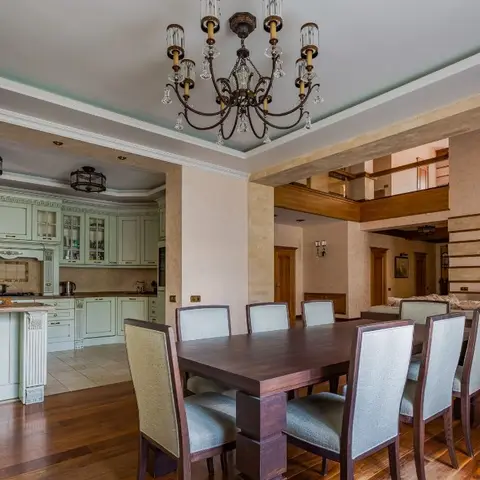 An open-plan kitchen, dining, and living space beneath a double-height mezzanine, with a crystal chandelier over the dining table and sage-green shaker kitchen
