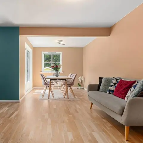 An open-plan living and dining space with a teal accent wall, peach walls and a light grey sofa with mixed-pattern cushions.