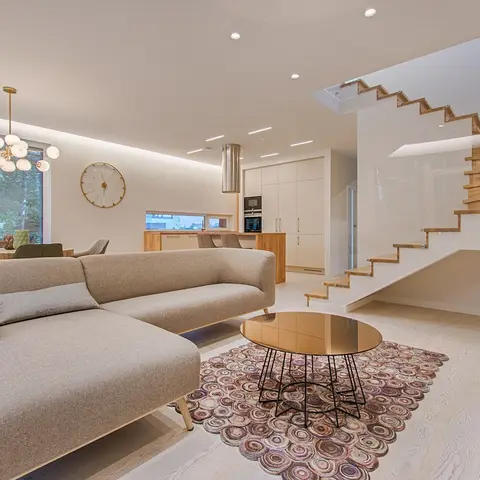 An open-plan living-dining-kitchen with a floating wood staircase, a beige sectional sofa on a circular patterned rug, a glass-top brass coffee table