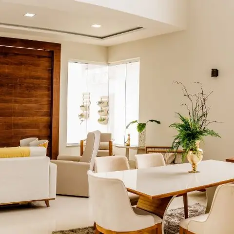 An open-plan living-dining space with a cream sectional and built-in TV shelving on one side, a wood dining table anchored by a large potted fern