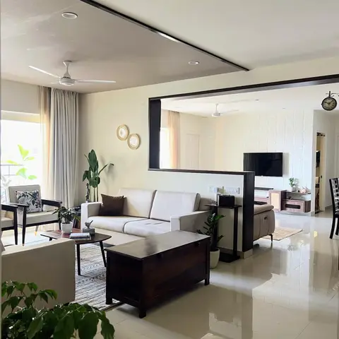 An open-plan living room with a beige sofa, a dark wood trunk-style coffee table, and a framed pass-through opening to a second lounge and TV area beyond.