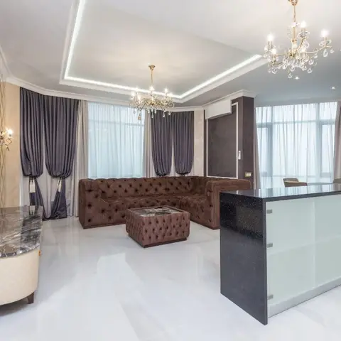An open-plan living space with a tufted brown leather chesterfield sofa, twin crystal chandeliers, floor-length curtains and a marble TV console
