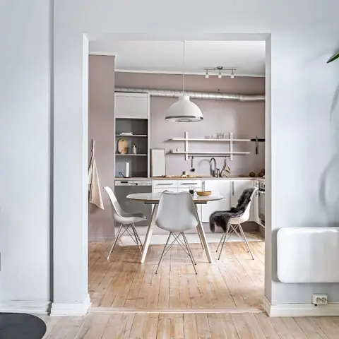 An open-plan Scandinavian space with a black wood-burning stove in the foreground and a pink-walled kitchen and round dining table visible through the doorway.