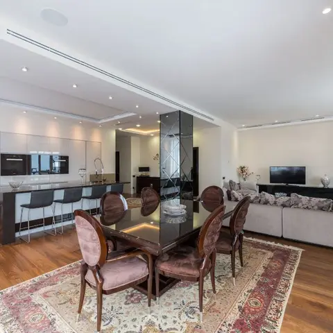 An open-plan space showing a kitchen island with bar stools, a carved-wood dining table on a Persian-style rug, and a living room sofa with TV console