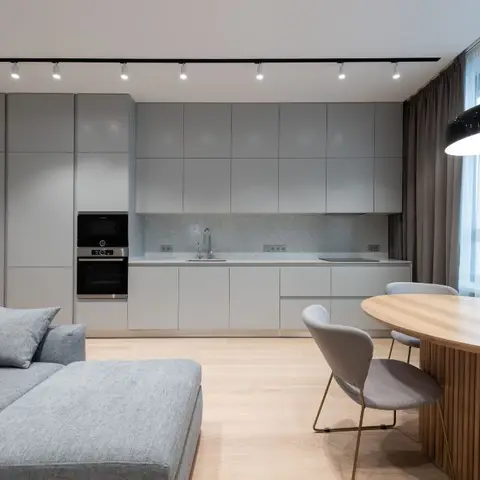 An open-plan space showing a flat-panel grey kitchen run, a round wood dining table beneath a black drum pendant light, and a grey sectional sofa corner