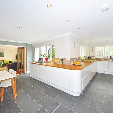 An open-plan white gloss U-shaped kitchen with a wood countertop and grey slate flooring leads directly to a small dining nook and living room beyond.
