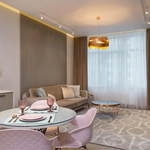 An open studio living-dining-kitchen area with blush pink dining chairs and glassware at a round table, a taupe velvet sofa, a copper ribbon pendant light