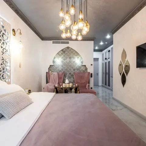 An opulent bedroom with a carved jaali-pattern headboard panel, a cluster bulb chandelier and a mosaic-tiled arched seating nook with velvet armchairs.