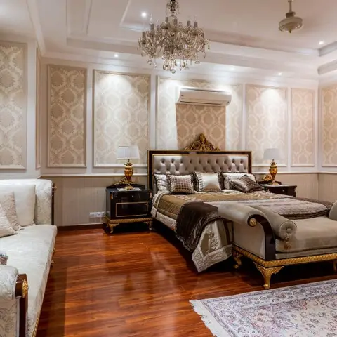 An opulent bedroom with a gilded tufted headboard, twin crystal chandeliers, cream damask wall panels and a matching chaise lounge at the foot of the bed.