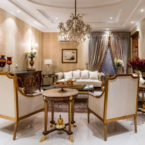 An ornate living room with gold damask wallpaper, a crystal chandelier, gilt-frame upholstered armchairs and sofa around a round marble coffee table