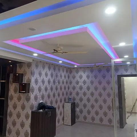 An unfurnished foyer with a multi-tier POP false ceiling lit by blue and pink cove LED strips and damask-patterned wallpapered walls