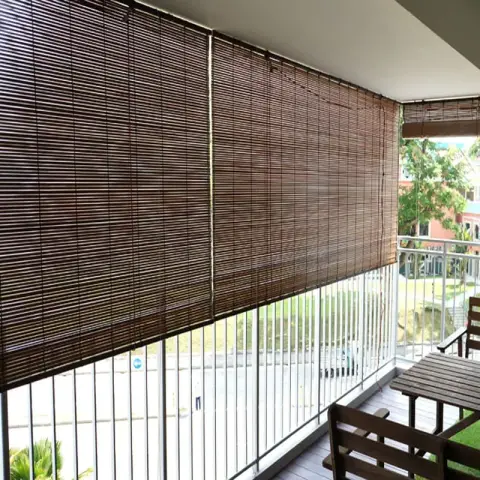 Apartment balcony screened with dark bamboo chick blinds rolled down over the railing, a small wooden outdoor table and chairs at the far end on grey decking.