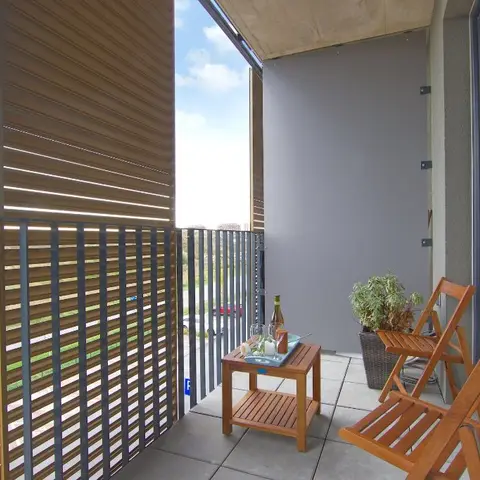 Apartment balcony with foldable wooden chairs and a small drinks table beside a potted plant, screened by louvred privacy panels.